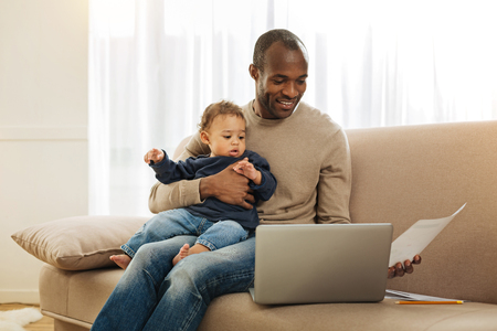 Paternal leave. Cheerful dark-eyed bearded afro-american man smiling and holding his son on his lap while working on the laptop and holding a sheet of paperの写真素材