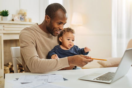 Favourite work. Good-looking cheerful dark-eyed bearded afro-american man smiling and working on the laptop and holding a sheet of paper and a fireplace in the backgroundの写真素材