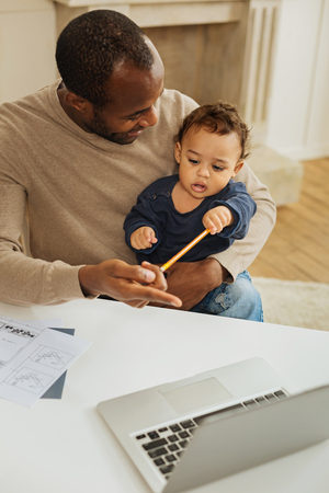 Working father. Handsome happy bearded afro-american man smiling and working on the laptop while a child sitting on his lapの写真素材