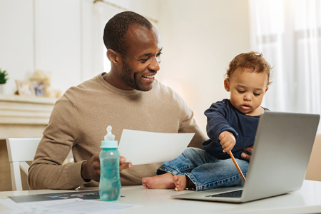Helping daddy. Good-looking happy bearded afro-american man smiling and watching his little son typing on the laptop and the bottle standing on the tableの写真素材