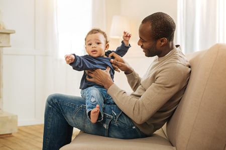 Amusing son. Good-looking happy afro-american man smiling and amusing his son while child is sitting...