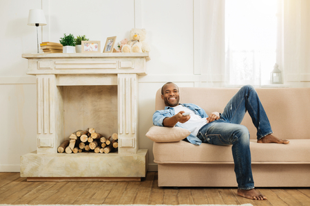 Relaxation. Handsome young dark-haired afro-american man grinning and holding remote control while relaxing on the couch and a fireplace near itの写真素材