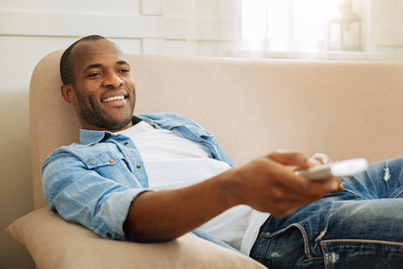 Sweet home. Attractive happy young dark-haired afro-american man grinning and holding remote control while lying on the couch and enjoying his weekendの写真素材