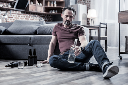 Pathetic man. Unhappy drunk cheerless man sitting on the floor and smoking a cigarette while holding a bottleの写真素材