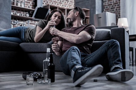 Alcohol addicted couple. Happy cheerful bearded man smiling and giving his wife a drink while sitting on the floorの写真素材
