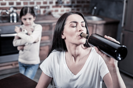 Careless mother. Sad unhappy young woman sitting at the table and holding a bottle while drinking alcoholの写真素材