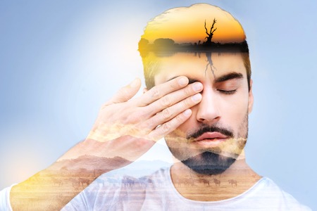 Absolutely calm. Nice handsome peaceful man closing his eye and covering one of them with his hand while imagining natureの写真素材