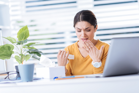 Sneeze. Beautiful young desperate woman feeling unwell while being ill and constantly sneezing when trying to work in her officeの写真素材