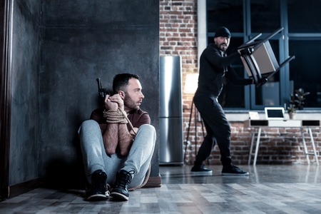 Hiding. Frightened shocked bearded man holding a gun and sitting with his hands tied while hiding from a criminal holding a chairの写真素材
