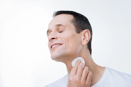 Skin care. Cheerful peaceful good looking man closing his eyes and cleaning his skin while using a cotton padの写真素材