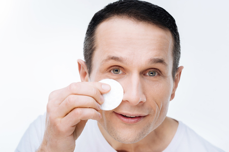 Facial care. Portrait of a nice handsome positive man holding a cotton pad and smiling while cleaning his face with a cotton padの写真素材