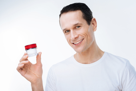 Facial cosmetics. Delighted positive nice man smiling and holding a bottle of facial cream while using itの写真素材
