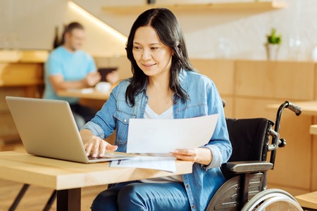 Freelancing. Beautiful happy dark-haired crippled woman sitting in a wheelchair and holding a sheet...