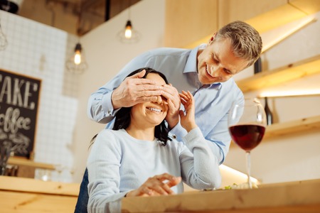Surprise. Attractive exuberant blond man closing his womans eyes and smiling while drinking wine in a cafeの写真素材