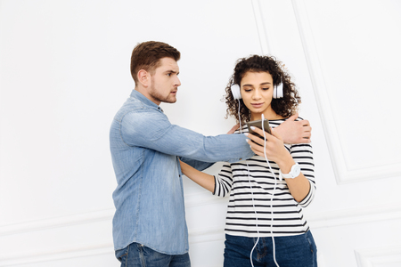 Look at me. Pretty woman with curly hair wearing headphones and looking at her gadget while choosing necessary songの写真素材