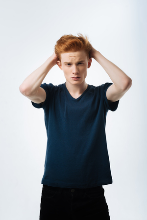 Hey. Attractive stony-faced red-haired young man touching his head with his hands and wearing a black t-shirt and looking seriouslyの写真素材