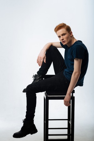 Stony-faced. Handsome concentrated red-haired young man sitting on the chair and putting one foot on it and wearing a black t-shirt and trousers and bootsの写真素材