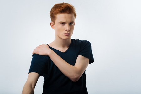 Staring. Handsome stern red-haired young man sitting and staring and having his hand on his shoulderの写真素材