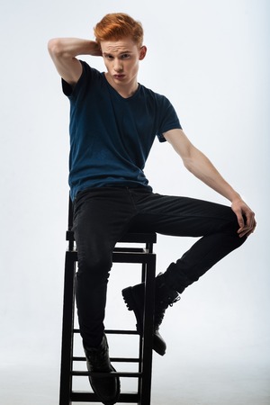 Pouting. Handsome determined red-headed young man sitting on the chair and pouting and having his hands on his neck and kneeの写真素材