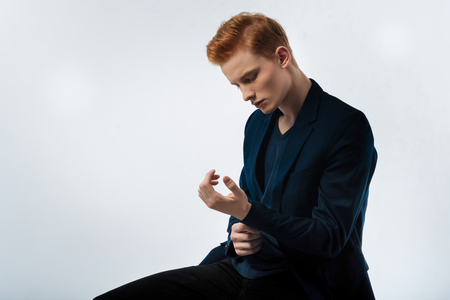 Thinking. Attractive earnest red-haired young man wearing a black jacket and touching his wrist and looking at itの写真素材