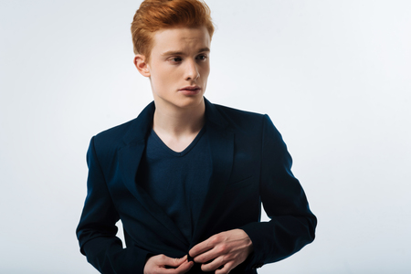 Stern. Good-looking earnest red-haired young man wearing a black jacket and buttoning it up and looking in the distanceの写真素材