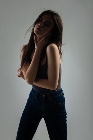 Nice. Beautiful stern dark-haired young woman staring and touching her neck and wearing a shirt and jeansの写真素材