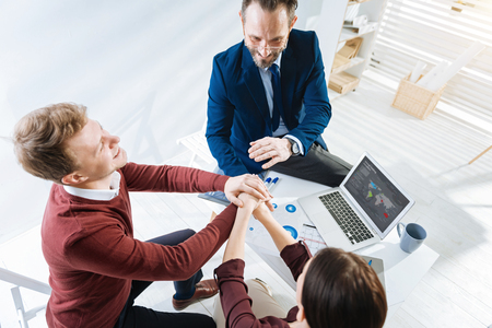 Team goal. Top view of happy cheerful three colleagues sitting while holding hands together and grinningの写真素材