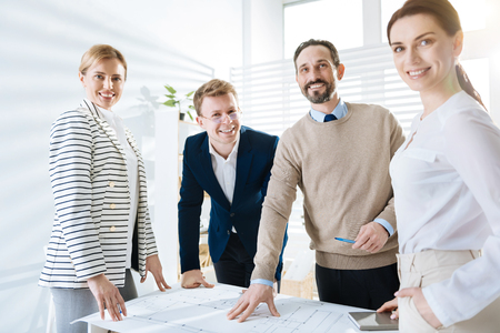 Ideal blueprint. Ambitious happy four  colleagues staying while laughing and gazing at the cameraの写真素材
