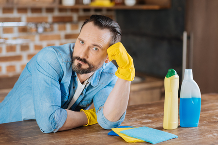 Boring cleaning. Attractive reflective male cleaner leaning on his hand while wearing gloves and gazing at the cameraの写真素材