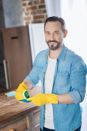 Less effort. Merry jolly male cleaner applying cleanser on the cleaning cloth while grinning and looking at the cameraの写真素材