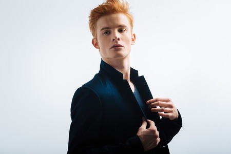 Serious. Handsome earnest red-headed young man wearing a black jacket and touching it and looking in the distanceの写真素材
