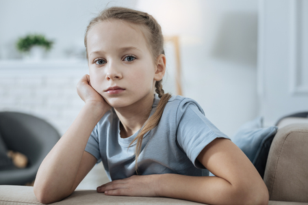 Sadness. Unhappy fair-haired blue-eyed little girl thinking and holding her head with her hand while...