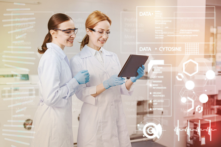 Cheerful young smiling doctor holding a modern tablet while her positive colleague standing by her side and looking at the screen of it together with herの写真素材