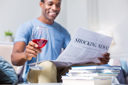 Alcoholic drink. Selective focus of a glass with red wine being in hands of a cheerful nice positive man while reading a newspaperの写真素材