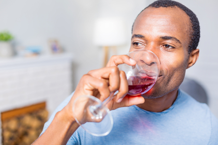Favourite drink. Portrait of a cheerful nice handsome man smiling and drinking wine while resting at homeの写真素材