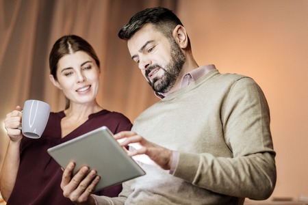 Relaxed atmosphere. Attentive bearded man standing in semi position and bowing head while looking at his gadgetの写真素材