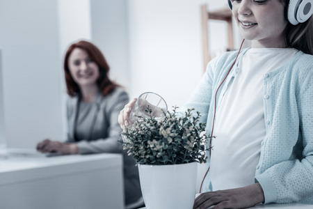 Proud parent. Careful girl in casual listening to music and watering a plant while her mother smiling and watching her in the background.の写真素材