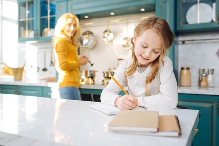 Beautiful happy fair-haired girl smiling and writing in her notebook while her mother standing in the backgroundの写真素材