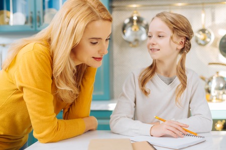 Beautiful exuberant fair-haired girl smiling and holding a pencil while her mother looking at her notebook and thinkingの写真素材
