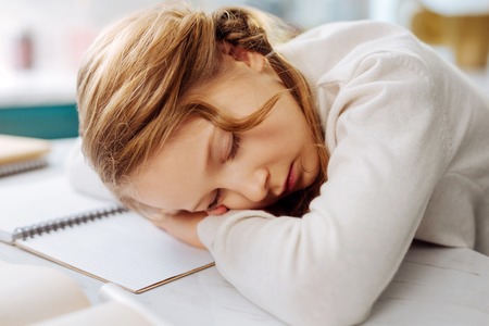 Pretty sweet fair-haired little schoolgirl sleeping over her books while sitting at the table and doing homeworkの写真素材
