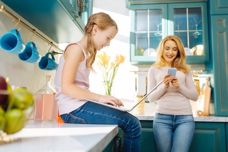Beautiful happy fair-haired girl smiling and working on the laptop and sitting on the table while her mother using her phoneの写真素材