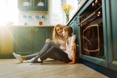 Beautiful content fair-haired mother and daughter smiling and sitting on the floor in the kitchen and embracingの写真素材