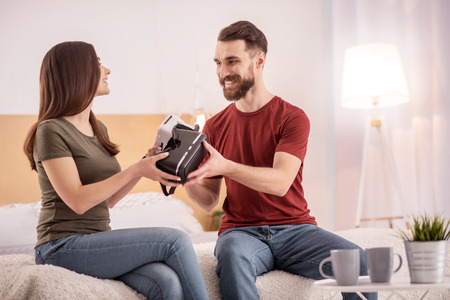 Here mine. Cute appealing young couple relaxing on the bad while stretching VR glasses to each other and drinking coffeeの写真素材