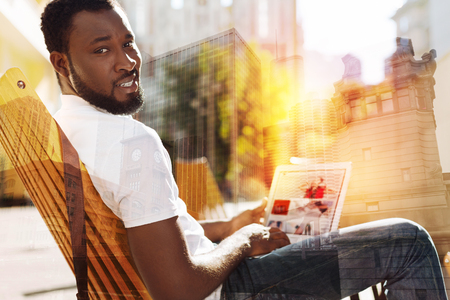 Busy man. Calm serious young man sitting in a wooden armchair with a modern convenient laptop on his knees and slightly frowning while turning his headの写真素材