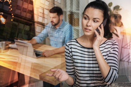 Beautiful young attentive woman talking on the phone and looking to the right while her enthusiastic serious colleague sitting behind her and workingの写真素材