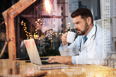 Clever experienced qualified doctor frowning while drinking coffee and looking attentively at the screen of his laptopの写真素材