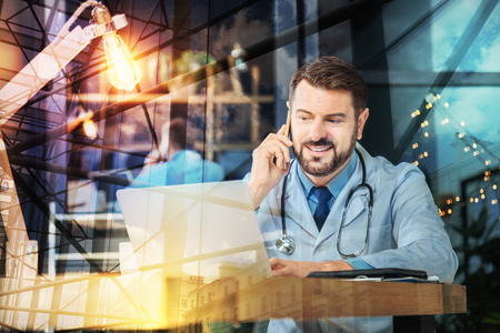 Positive conversation. Cheerful emotional handsome doctor looking at the screen of his convenient laptop and smiling while having a pleasant phone talkの写真素材
