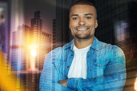 Cheerful smile. Young calm cheerful man standing alone with his arms crossed and smiling while waiting for his close friendの写真素材