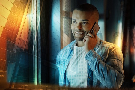 Phone talk. Positive friendly young man looking glad while standing near the window and having a pleasant phone talkの写真素材