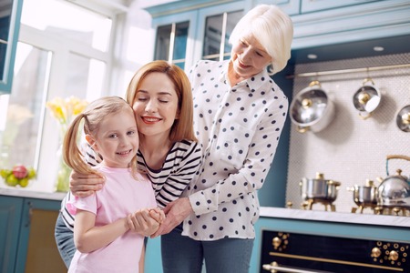 Happy family. Pleasant little girl standing in the kitchen and being hugged by her mother and...
